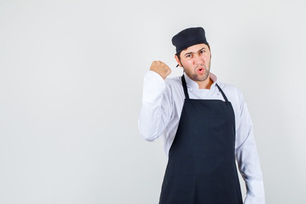 Male chef showing up clenched fist in uniform, apron and looking indignant. front view.