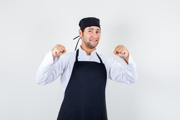 Male chef showing winner gesture in uniform, apron and looking happy , front view.