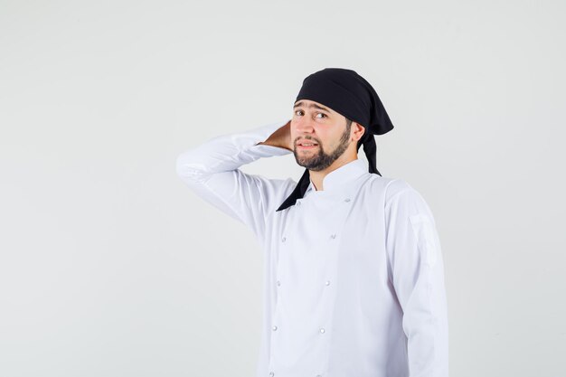 Male chef standing with hand behind head in white uniform and looking irresolute , front view.