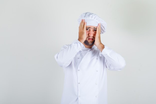 Male chef standing with head in hands in white uniform and looking confused.