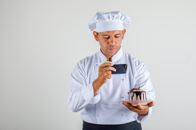 Мужской шеф-повар фотографирует десерт торт в форме, фартук и шляпу