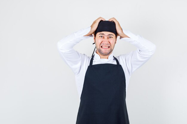 Male chef in uniform, apron clenching teeth with hands on head and looking stressful , front view.