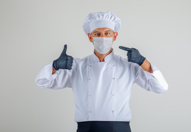 Free photo male chef in uniform, apron and hat showing mask and thumbs up and looking careful