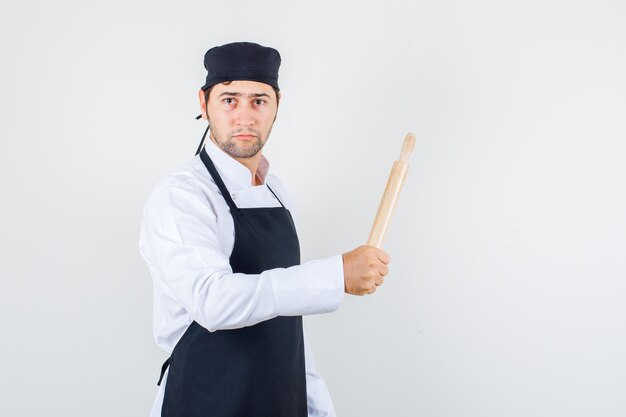 Шеф-повар-мужчина в униформе, агрессивно держит скалку в фартуке и смотрит строго, вид спереди.