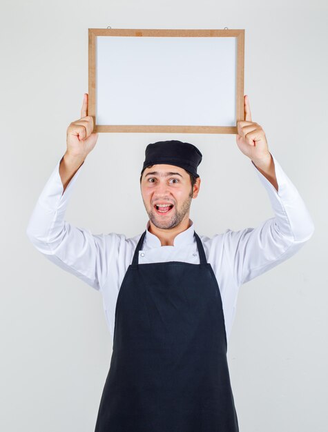 Male chef in uniform, apron holding white board over head and looking cheery , front view.