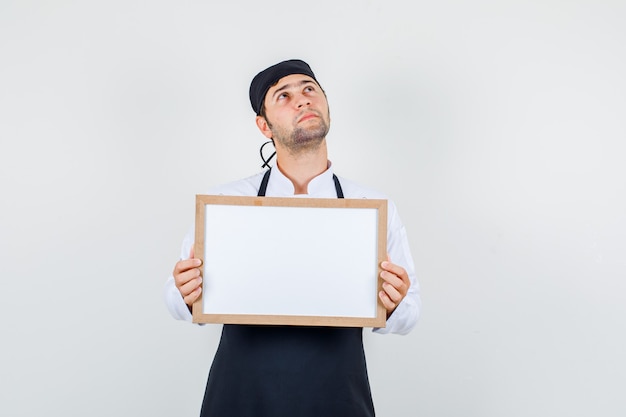 Male chef in uniform, apron holding white board and looking pensive , front view.
