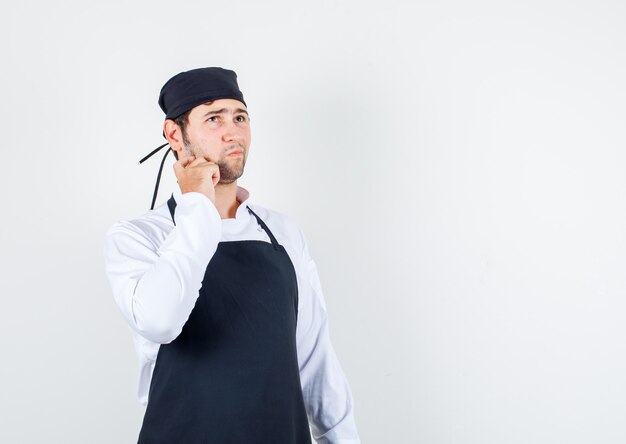 Foto gratuita chef maschio in uniforme, grembiule alzando lo sguardo con il dito sulla guancia e guardando pensieroso, vista frontale.