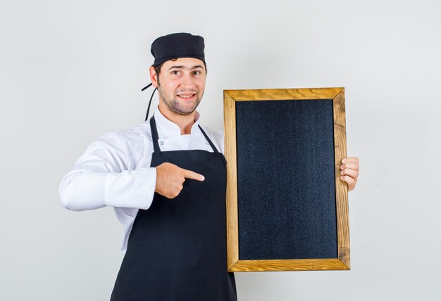 Male chef in uniform, apron pointing at blackboard and looking cheery , front view.