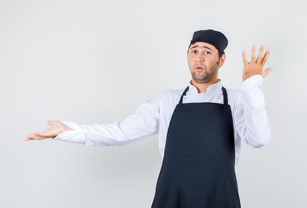 Шеф-повар-мужчина в униформе, в фартуке разводит ладонь, поднимает руку и выглядит смущенным, вид спереди.
