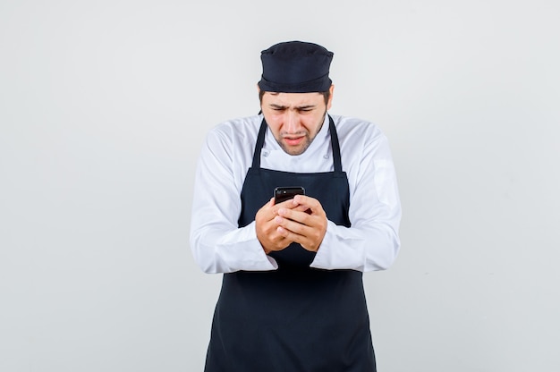 Male chef in uniform, apron using smartphone and looking puzzled , front view.