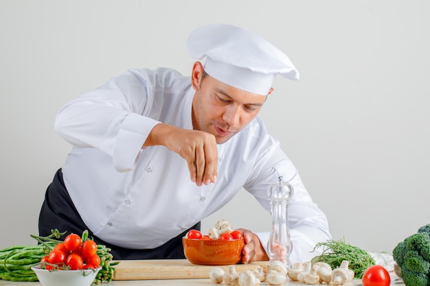 Male chef in uniform, hat and apron adding spice into food in kitchen