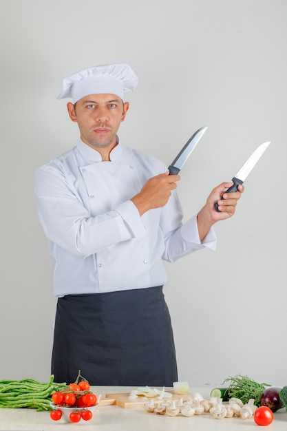 Male chef in uniform, hat and apron holding metal knives in kitchen