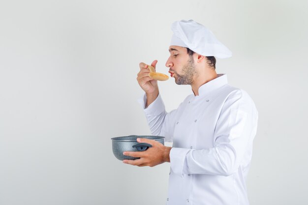 Male chef in white uniform blowing on hot soup and holding saucepan