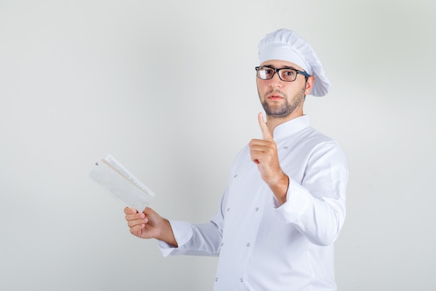 Male chef in white uniform, glasses holding book with waiting gesture