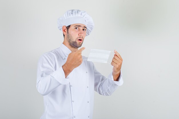 Мужчина-шеф-повар в белой форме держит медицинскую маску и выглядит нерешительно