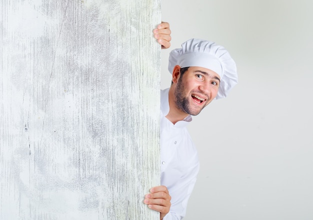 Chef maschio in uniforme bianca guardando da dietro la tavola di legno e guardando allegro