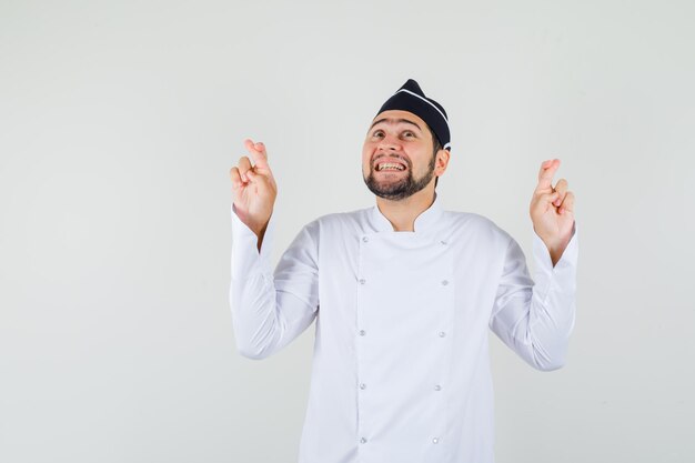 Male chef in white uniform looking up with crossed fingers and looking merry , front view.