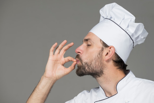 Male chef in white uniform making tasty sign against gray background