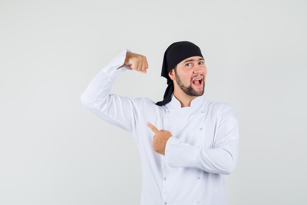 Male chef in white uniform pointing finger at his arm muscles and looking confident , front view.