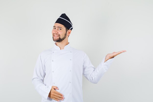 Male chef in white uniform raising his hand like showing something and looking pleased , front view.