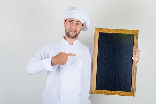 Male chef in white uniform showing blackboard with finger and looking positive 