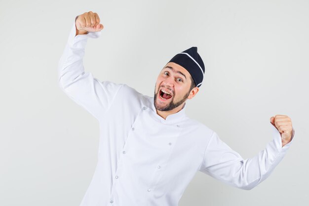 Chef maschio in uniforme bianca che mostra il gesto del vincitore e sembra allegro, vista frontale.