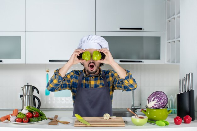 Chef maschio con verdure fresche e cucinare con utensili da cucina e tenere i peperoni verdi tagliati che si coprono gli occhi nella cucina bianca