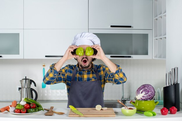 Шеф-повар-мужчина со свежими овощами и готовкой с кухонными принадлежностями, прикрывая глаза нарезанным зеленым перцем на белой кухне
