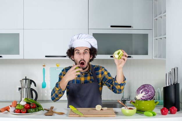 Шеф-повар-мужчина со свежими овощами и приготовлением пищи с кухонными принадлежностями и держит нарезанный зеленый перец, чувствуя себя удивленным на белой кухне