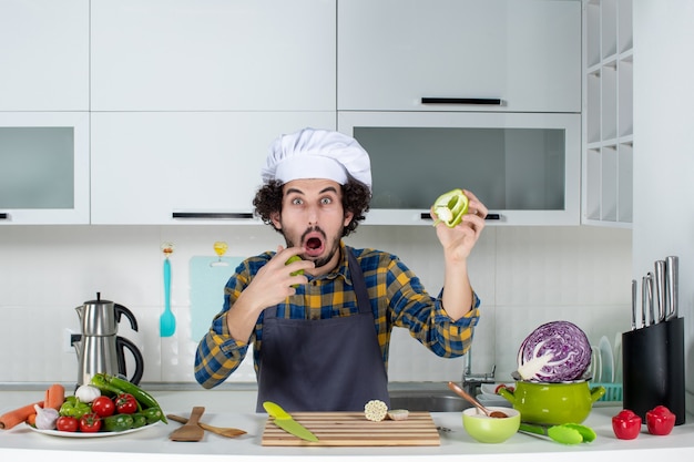 Male chef with fresh vegetables and cooking with kitchen tools and holding the cut green peppers feeling surprised in the white kitchen