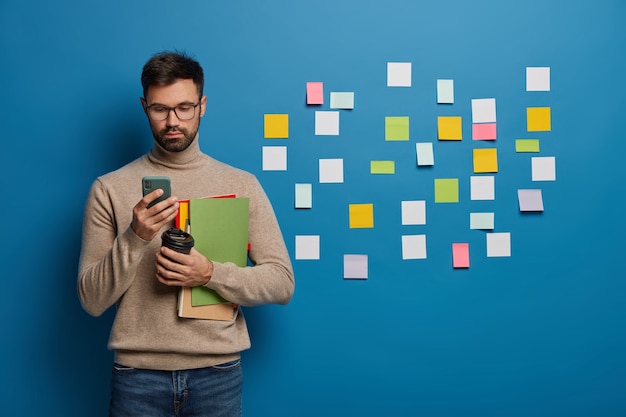 Foto gratuita studente universitario maschio utilizza il telefono cellulare per chattare online, beve caffè da asporto, tiene blocchetti per appunti o libri di testo, si prepara per la lezione, sta dietro il muro blu con molte note adesive