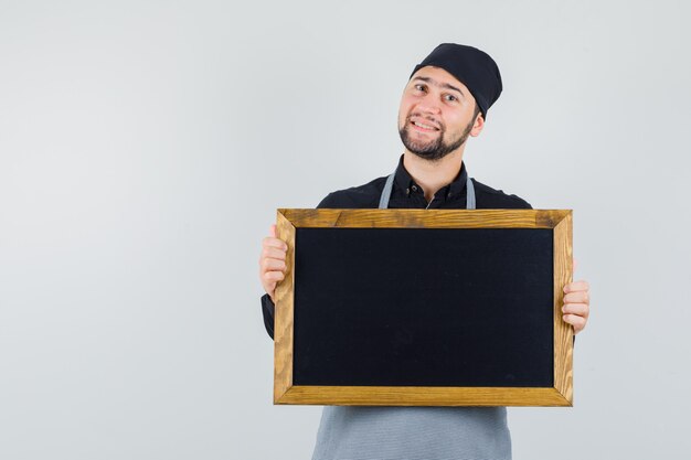 Male cook holding blackboard in shirt, apron and looking merry. front view.