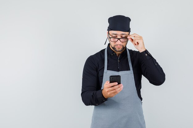 シャツ、エプロン、忙しそうに見えるメガネを通して携帯電話を見ている男性料理人。正面図。