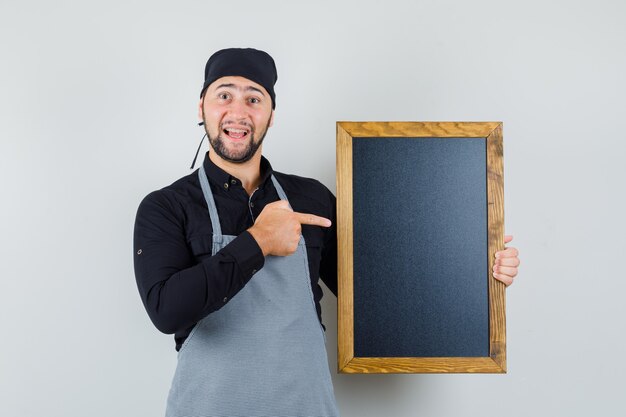 Male cook pointing at blackboard in shirt, apron and looking happy. front view.