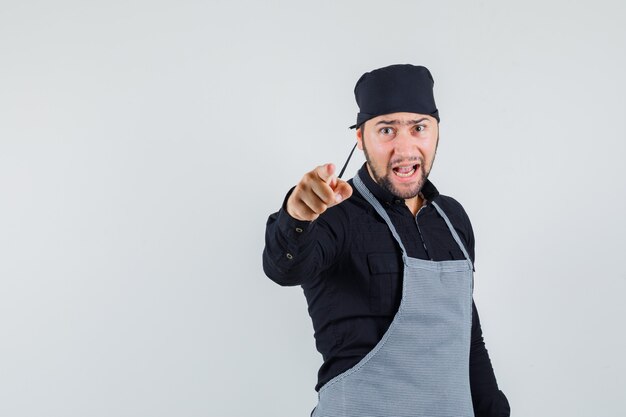 Male cook pointing at camera in shirt, apron and looking nervous , front view.