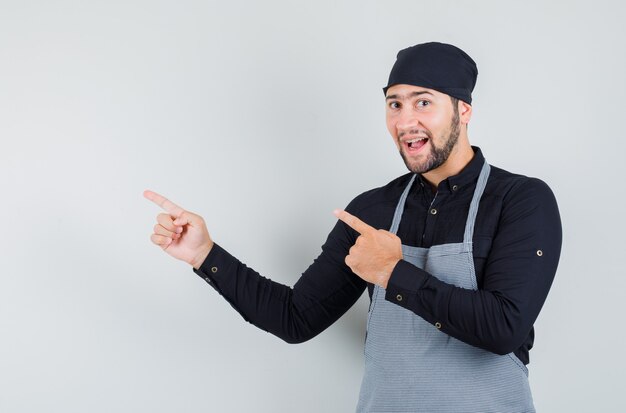 Male cook in shirt, apron pointing away and looking cheerful , front view.