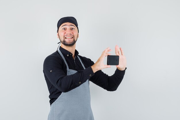 Male cook taking photo on mobile phone in shirt, apron and looking cheery. front view.