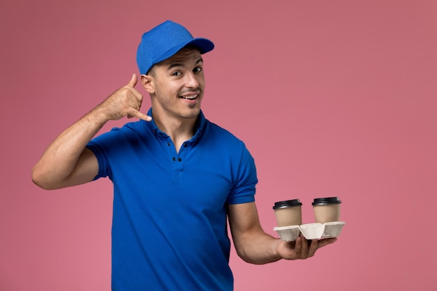 male courier in blue uniform holding brown coffee cups and posing on pink, worker uniform service delivery
