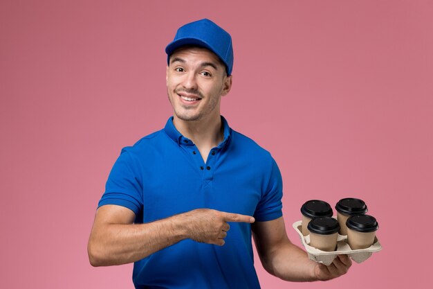 male courier in blue uniform holding brown coffee cups and posing on pink, worker uniform service delivery