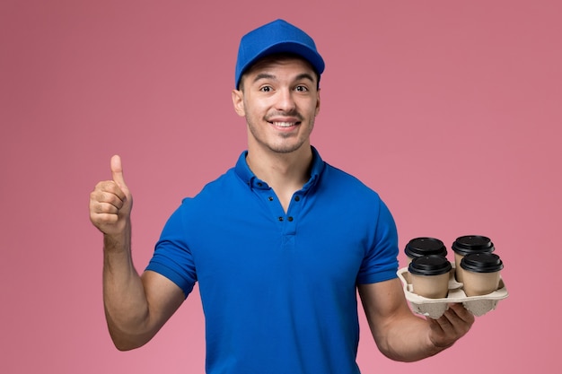 male courier in blue uniform holding brown coffee cups and smiling on pink, worker uniform service delivery