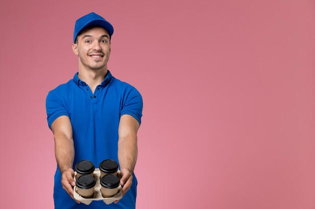 курьер-мужчина в синей форме держит коричневые кофейные чашки и улыбается на розовом, работник службы доставки униформы