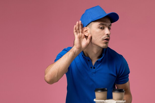 male courier in blue uniform holding brown coffee cups trying to hear on pink, worker uniform service delivery