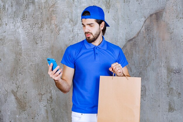 Male courier in blue uniform holding a cardboard shopping bag and talking to the phone.