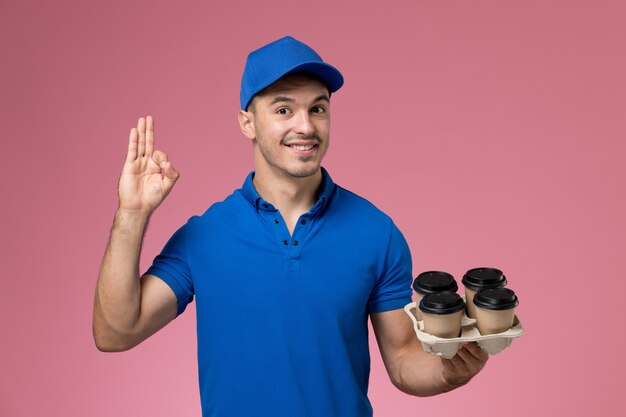 курьер-мужчина в синей форме держит кофейные чашки и позирует на розовом, служба доставки униформы