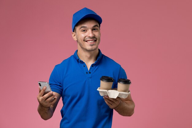 male courier in blue uniform holding coffee cups using his phone on pink, job worker uniform service delivery