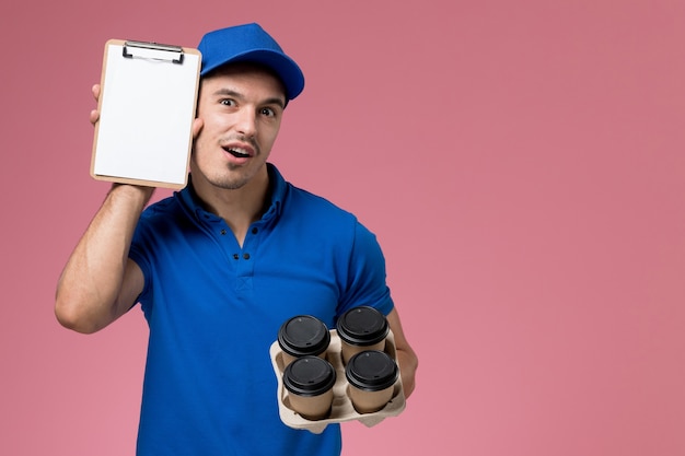 Free photo male courier in blue uniform holding coffee cups with notepad on pink, job uniform service delivery