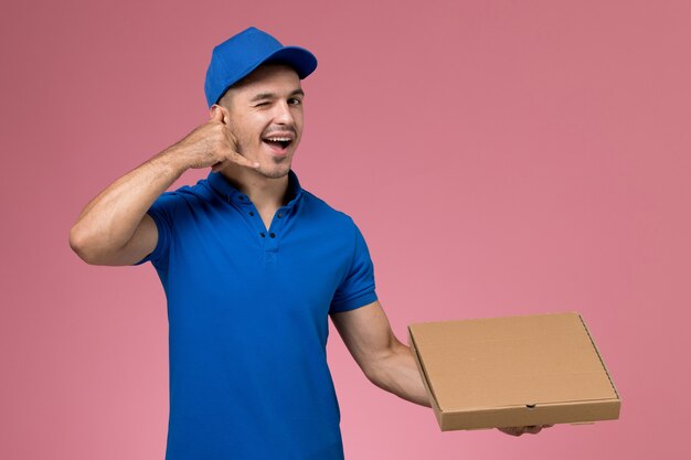 male courier in blue uniform holding delivery box of food posing on pink, worker uniform service delivery