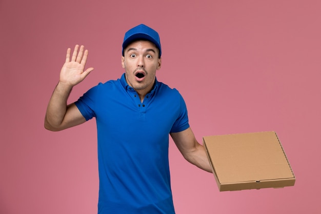 Male courier in blue uniform holding delivery box of food with surprised expression on pink, job worker uniform service delivery