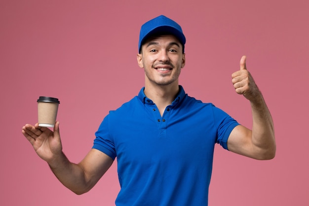 мужчина-курьер в синей форме держит чашку кофе с доставкой, улыбаясь и позирует на розовой, униформе работник службы доставки