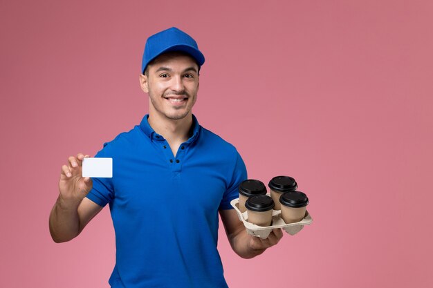 male courier in blue uniform holding delivery coffee cups and card smiling on pink, uniform service job delivery